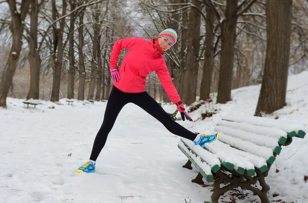 Winter running in park: happy woman runner warming up and exercising before jogging in snow, outdoor sport and fitness concept