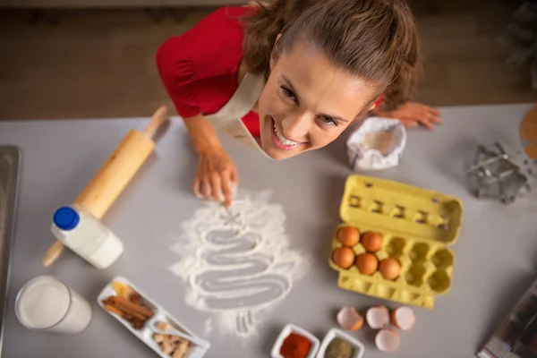 Happy young housewife drawing christmas tree with flour on table
