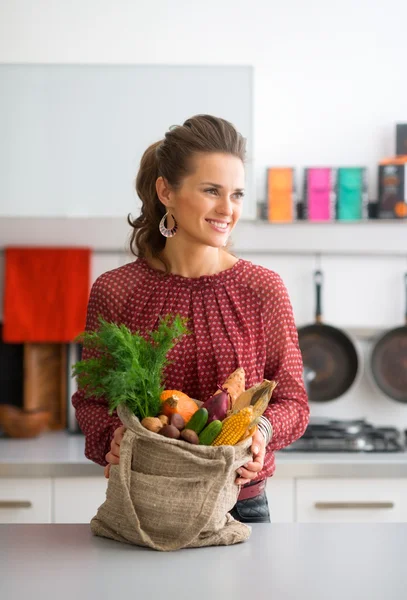 Smiling woman holding bag of autumn vegetables in kitchen