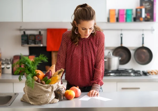 Woman in kitchen reading shopping list on counter with shopping