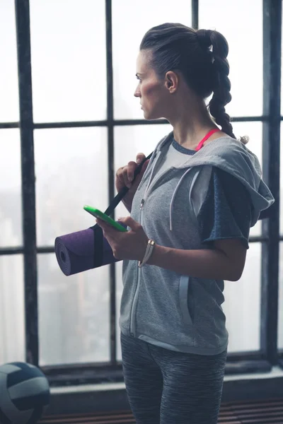 Woman in workout gear holding phone and yoga mat in loft gym