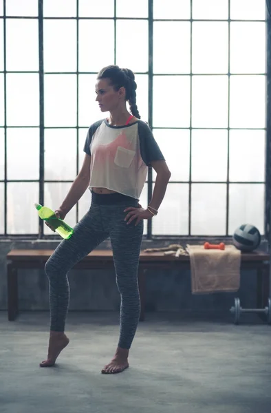 Woman standing in loft gym resting water bottle against leg