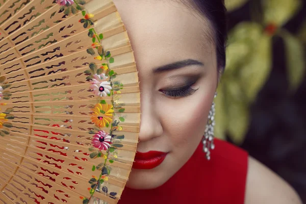 Beautiful asian female portrait with red dress and lips sitting on the green floral background holding wooden fan with flower pattern near her face