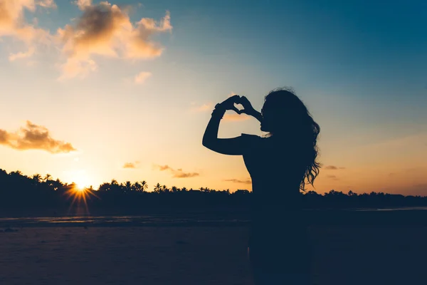 Silhouette of sexy brunette in fashion swimwear making hand gestures, showing love at sunset