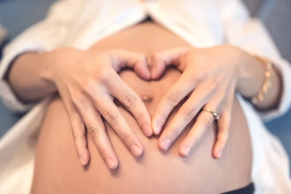 Pregnant young woman holding fingers in a heart shape, tounching belly on baby bump and loving the baby. Mother love concept.