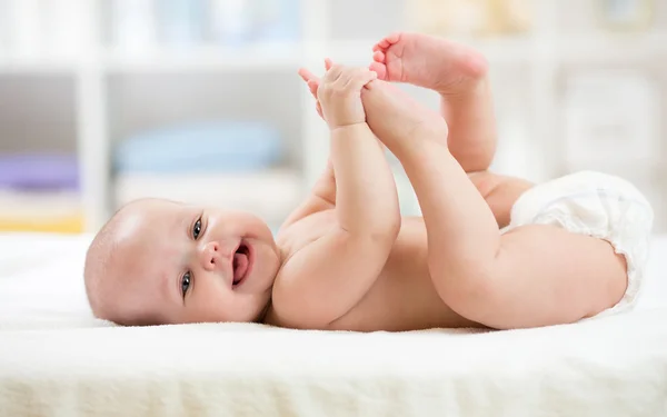 Funny little baby wearing a diaper playing on bed in nursery room. Happy child after bath or shower. Infant kid diaper change and skin care. Smiling kid playing with his feet.