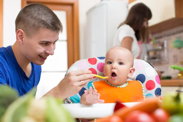 Young dad feeding his baby and mom cooking at kitchen