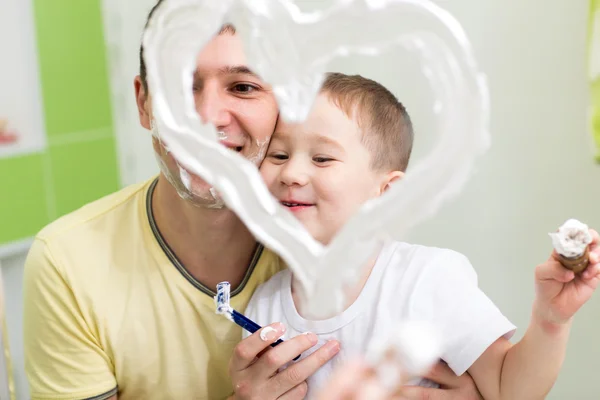 Father and child son drawing heart shape on mirror with shaving foam playing in bathroom
