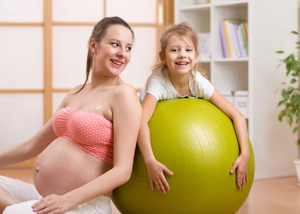 Pregnant mother and daughter doing gymnastics and smiling while sitting on floor with fitness ball