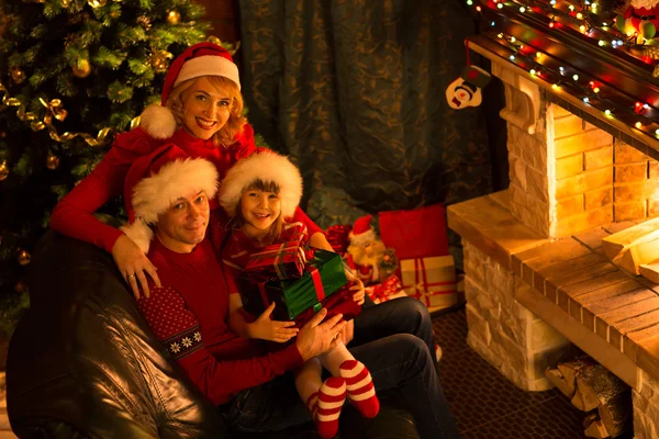 Happy family of three persons in red hats with gifts sitting at Christmas tree near fireplace