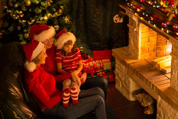 Family - mother, father and their little daughter sitting by a fireplace in a cozy dark living room on Christmas eve