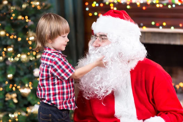 Surprised little boy looks at fake Santa Claus with fake beard sitting opposite Christmas tree