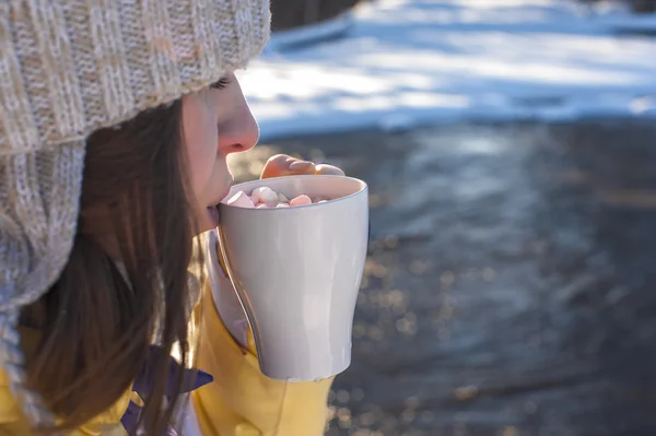 Woman holds white cup of coffee with marshmallows. Sunny winter day. Girl drinking coffee with marshmallows next to the river.
