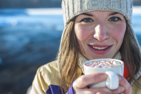 Smiling Woman holds white cup of coffee with marshmallows. Sunny winter day. Girl drinking coffee with marshmallows next to the river.