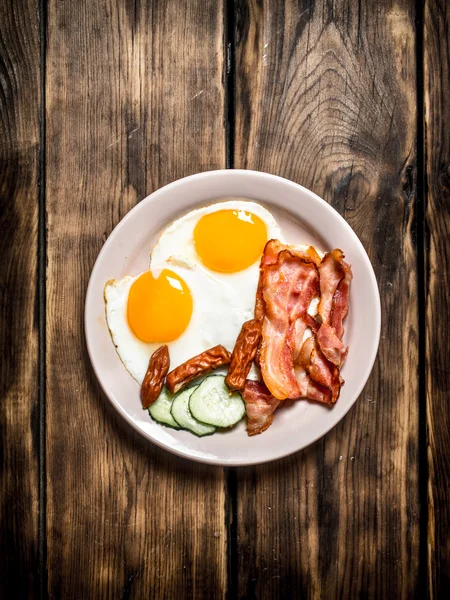 Plate of fried eggs, bacon , cucumber and smoked sausages.