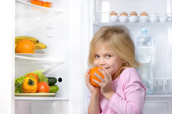 Little girl holding orange near open fridge
