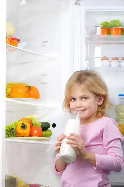 Little cute girl holding bottle of milk near open fridge