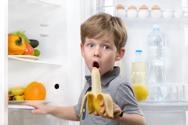 Little cute boy eating banana near open fridge