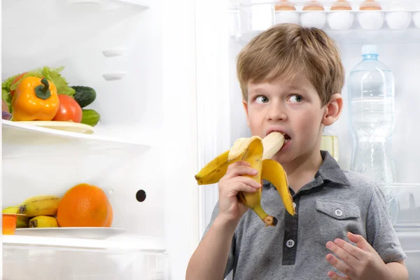 Cute boy eating banana near open fridge