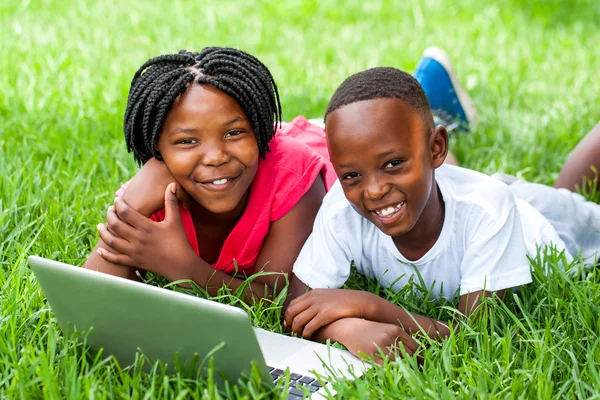 Two african kids laying on grass with laptop.