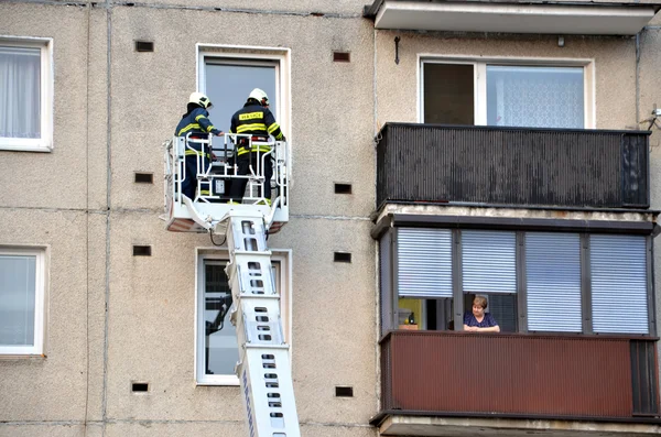 Two firefighters into telescopic boom basket of fire truck try to get to the flat balcony. Old woman is watching them from next balcony.