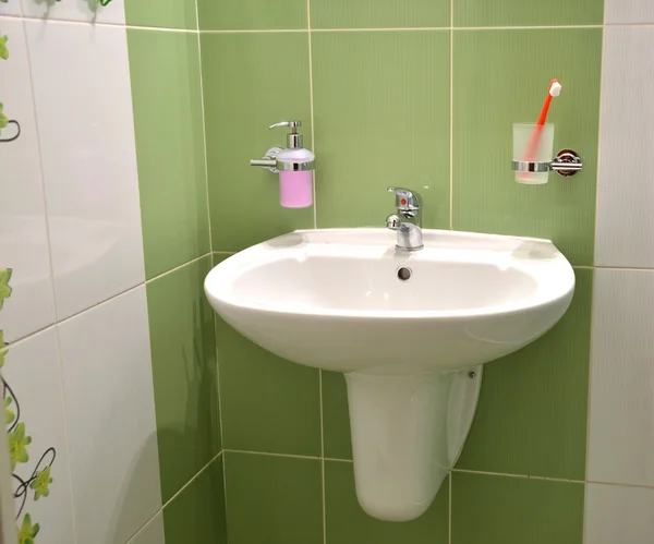 White wash basin, soap container and toothbrush in glass in white green tiled bathroom