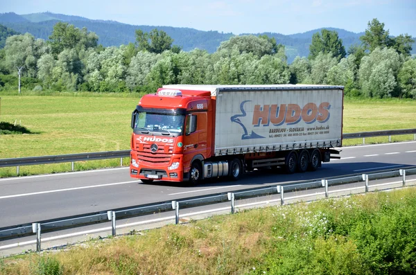 Red moving Mercedes-Benz Actros truck coupled with semi-trailer located on slovak D1 highway surrounded by green field and trees.