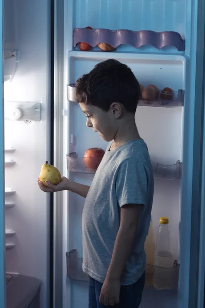 Child looking at a pear in front of the refrigerator in the midd