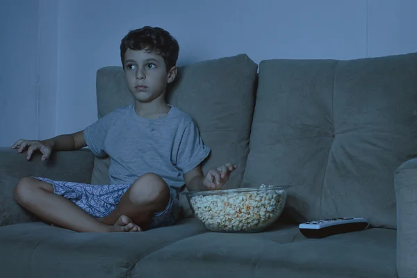 Small boy on the couch watching TV and eating popcorn at night i