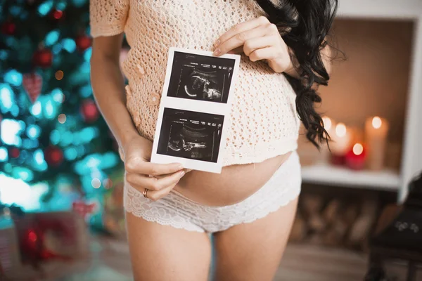 Pregnant woman holding ultrasound examination picture in front of her round belly, xmas
