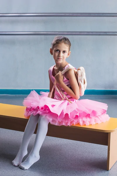 Beautiful little ballerina in pink dress for dancing sitting on the bench with pointe shoes in ballet studio