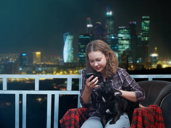 Girl with smart phone and a dog in his arms on the background of the city at night and skyscrapers
