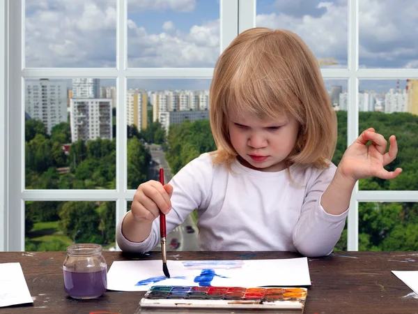 The child draws the table. Girl little blonde. Multicolored paint brush with blue. The window in the building. Outside, the city, the green trees, panoramic view