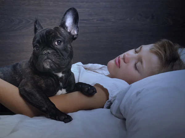 Young girl sleeping on the bed. Dog - French Bulldog. Evening or night, in a dark room