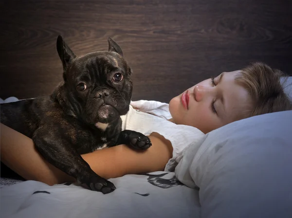 Young girl sleeping on the bed. Dog - French Bulldog. Evening or night, in a dark room