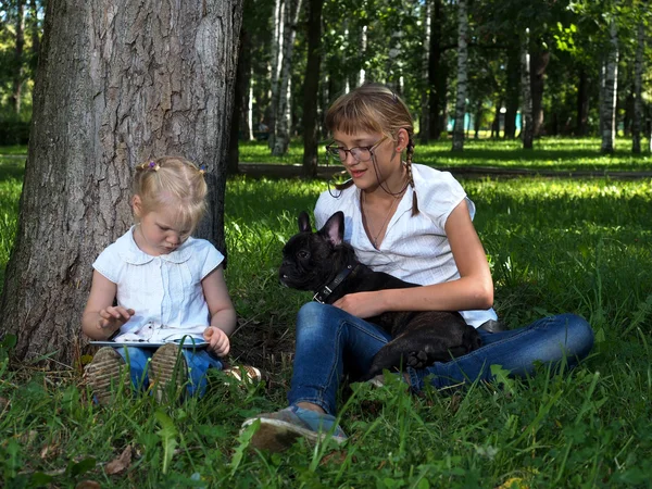 Two girls - adolescent and child sit in the park with tablet computer and dog