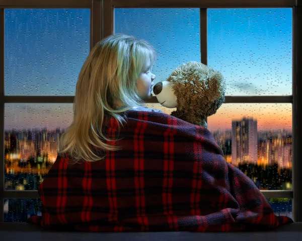 Little girl talking with a toy bear in the window. Outside, the late afternoon, the panorama of the city, rain. Raindrops on glass window