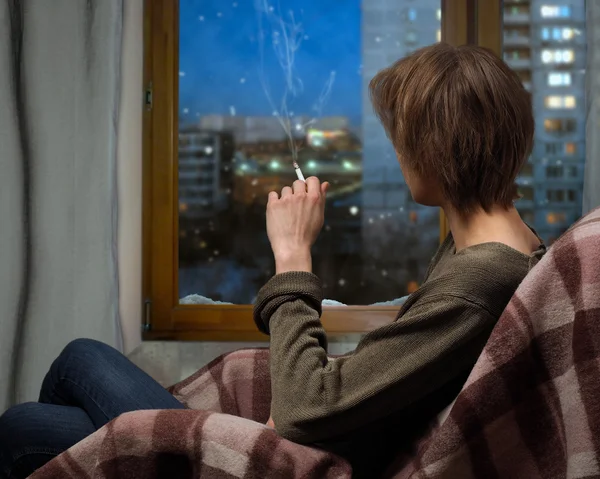 A woman smokes a cigarette while sitting in a chair. Outside the window, the snow falls. Evening, city block, the windows of houses. A woman looks out the window. Warm blanket