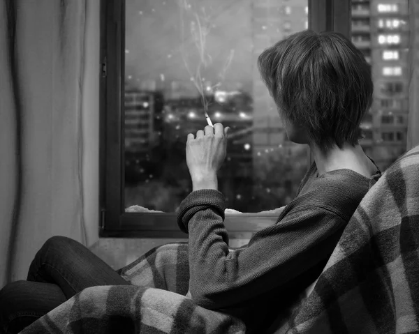 A woman smokes a cigarette while sitting in a chair. Outside the window, the snow falls. Evening, city block, the windows of houses. A woman looks out the window. A warm blanket. Black and white