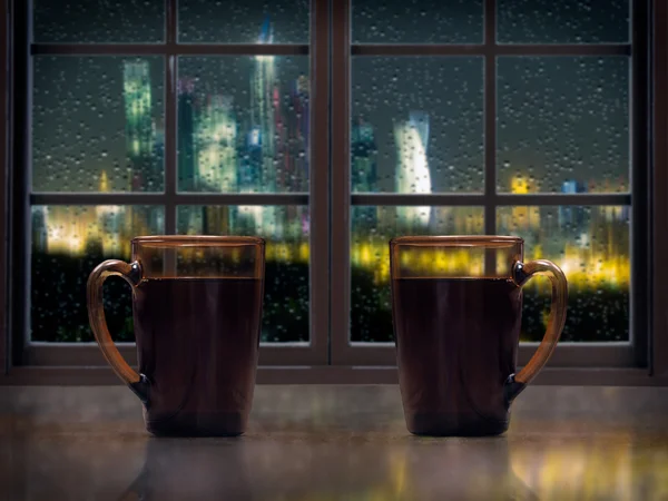 Two mugs with a hot drink - tea or coffee on the window sill of the window. Outside, the night, the city lights, rain, drops on the glass