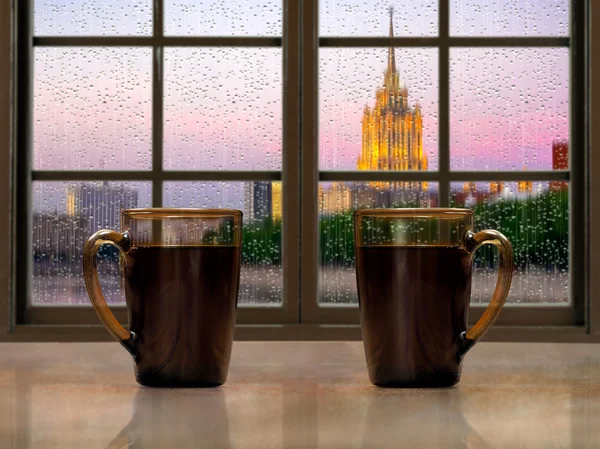 Two mugs with a hot drink - tea or coffee on the window sill of the window. During the early morning, the city, the rain. Concept - good morning alertness, awakening together