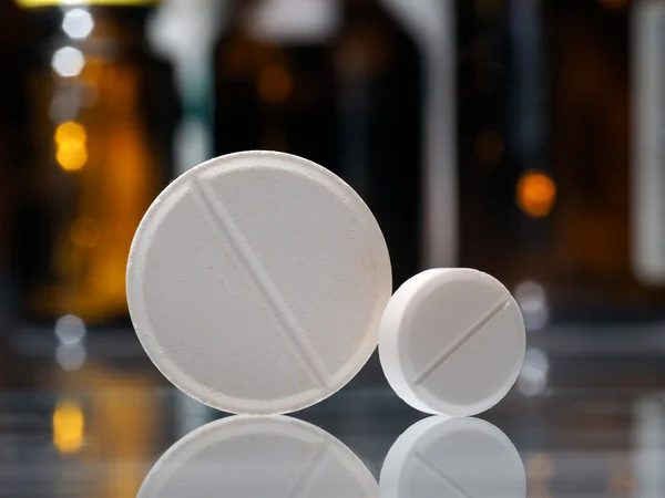 Two tablets. White pills, big and small. Background - dark bottles with medicine
