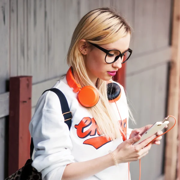 Girl with orange headphones looking for awesome track in her phone. Street shoot, music energy, lifestyle, european or american style, clear sound, sunshine, outdoors