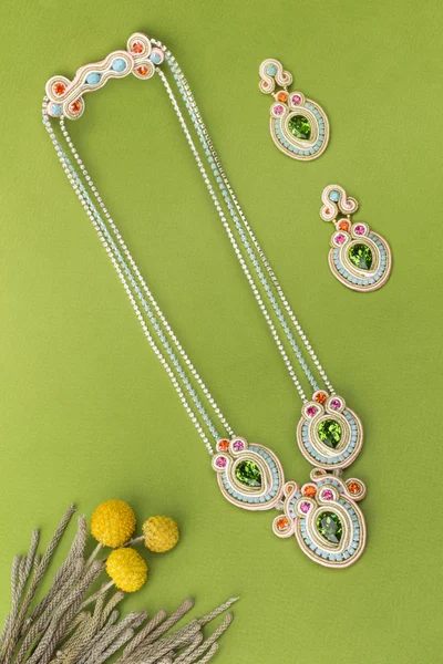 Soutache bijouterie set colorful earrings and necklaces with green purple cyan and pink crystals on green background with grass stems and yellow flowers
