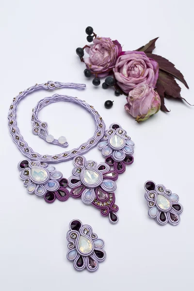 Soutache bijouterie set purple earrings and necklaces with white purple and pink crystals on white background with small roses black berries and dark red leaves
