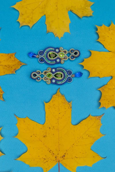 Soutache bijouterie blue earrings with green cyan and blue crystals on cyan background with yellow autumn maple leaves