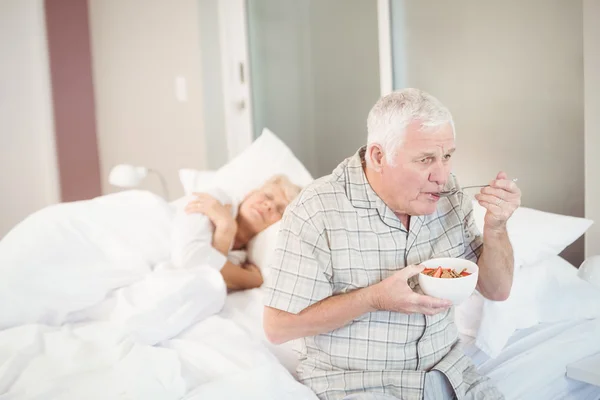Senior man eating salad by sleeping wife