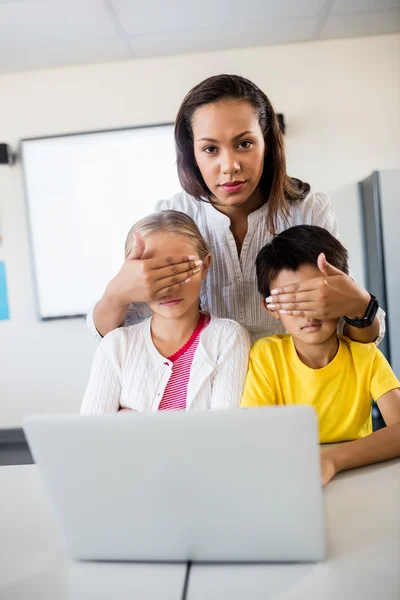 Teacher covering pupils eyes in front of computer and looking at