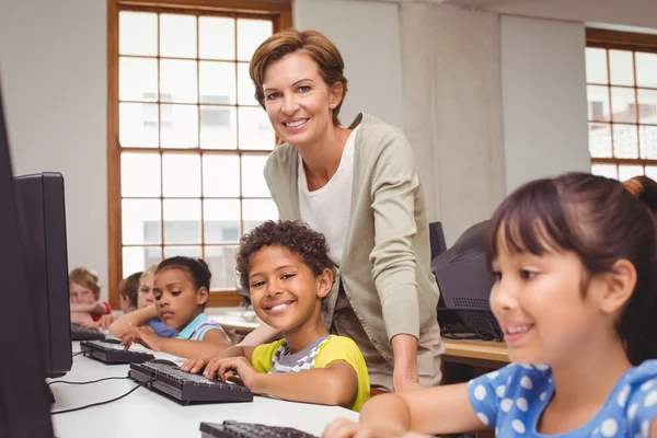 Cute pupil in computer class with teacher smiling at camera