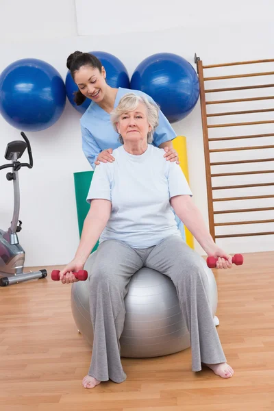 Therapist helping senior woman fit dumbbells on exercise ball